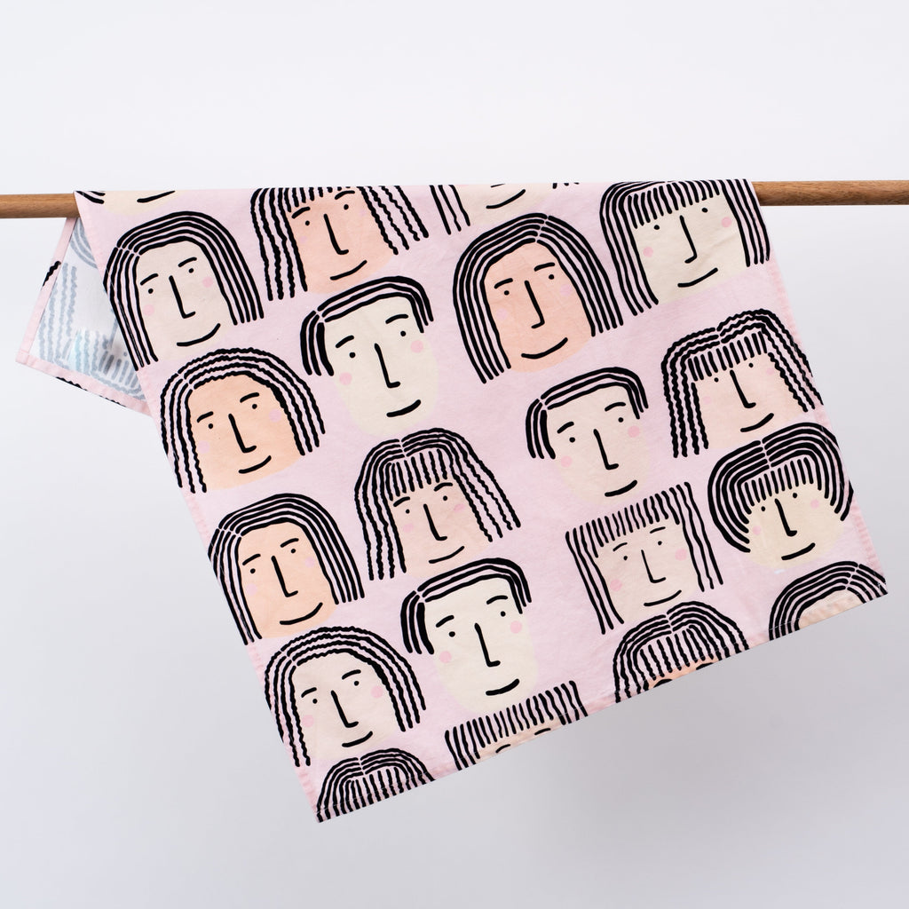 Fab faces tea towel. Different diverse faces, with different features, on a pink background.