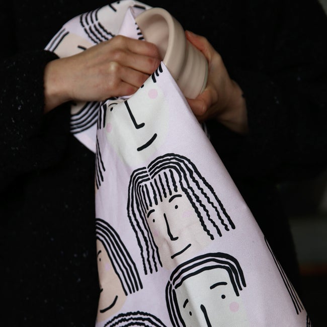 Fab faces tea towel. Different diverse faces, with different features, on a pink background. This picture shows the towel being used. 