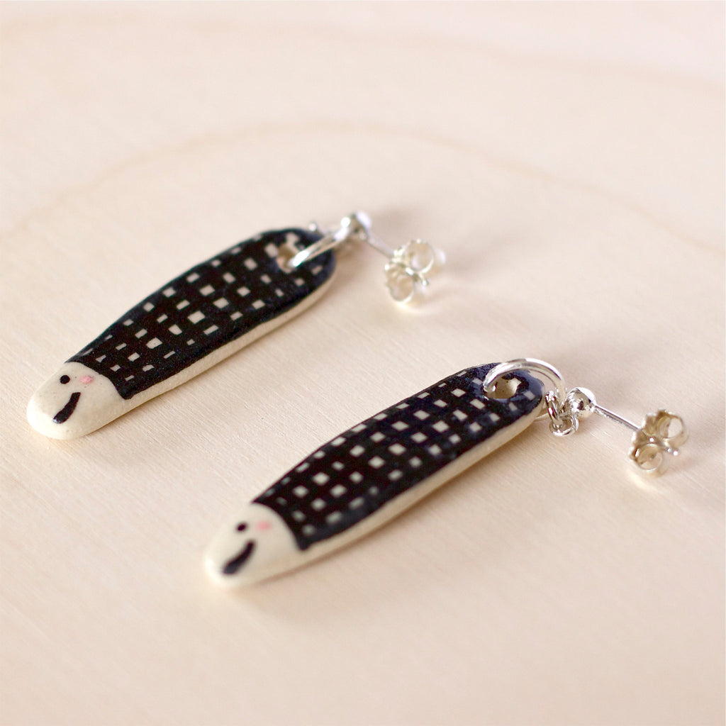 Flat view of a Pair of handmade ceramic earrings shaped like fish with a black check pattern and tiny white faces, finished with silver stud backings. Designed by shuh.