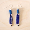 Top down view of a pair of handmade ceramic earrings shaped like fish with a navy blue check pattern and small face details, finished with silver stud hooks and blue fabric wrapping . Designed by shuh.