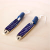Pair of handmade ceramic earrings shaped like fish with a navy blue check pattern and small face details, finished with silver stud hooks and blue fabric wrapping . Designed by shuh.