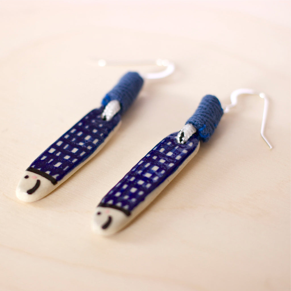Pair of handmade ceramic earrings shaped like fish with a navy blue check pattern and small face details, finished with silver stud hooks and blue fabric wrapping . Designed by shuh.
