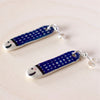 Flat view of a pair of handmade ceramic earrings shaped like fish with a navy blue check pattern and small face details, finished with silver stud posts. Designed by shuh.