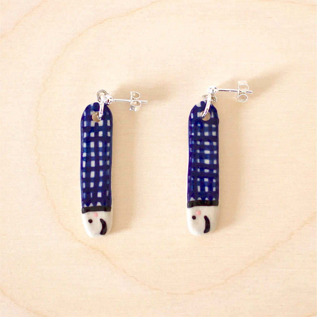 Pair of handmade ceramic earrings shaped like fish with a navy blue check pattern and small face details, finished with silver stud posts. Designed by shuh.