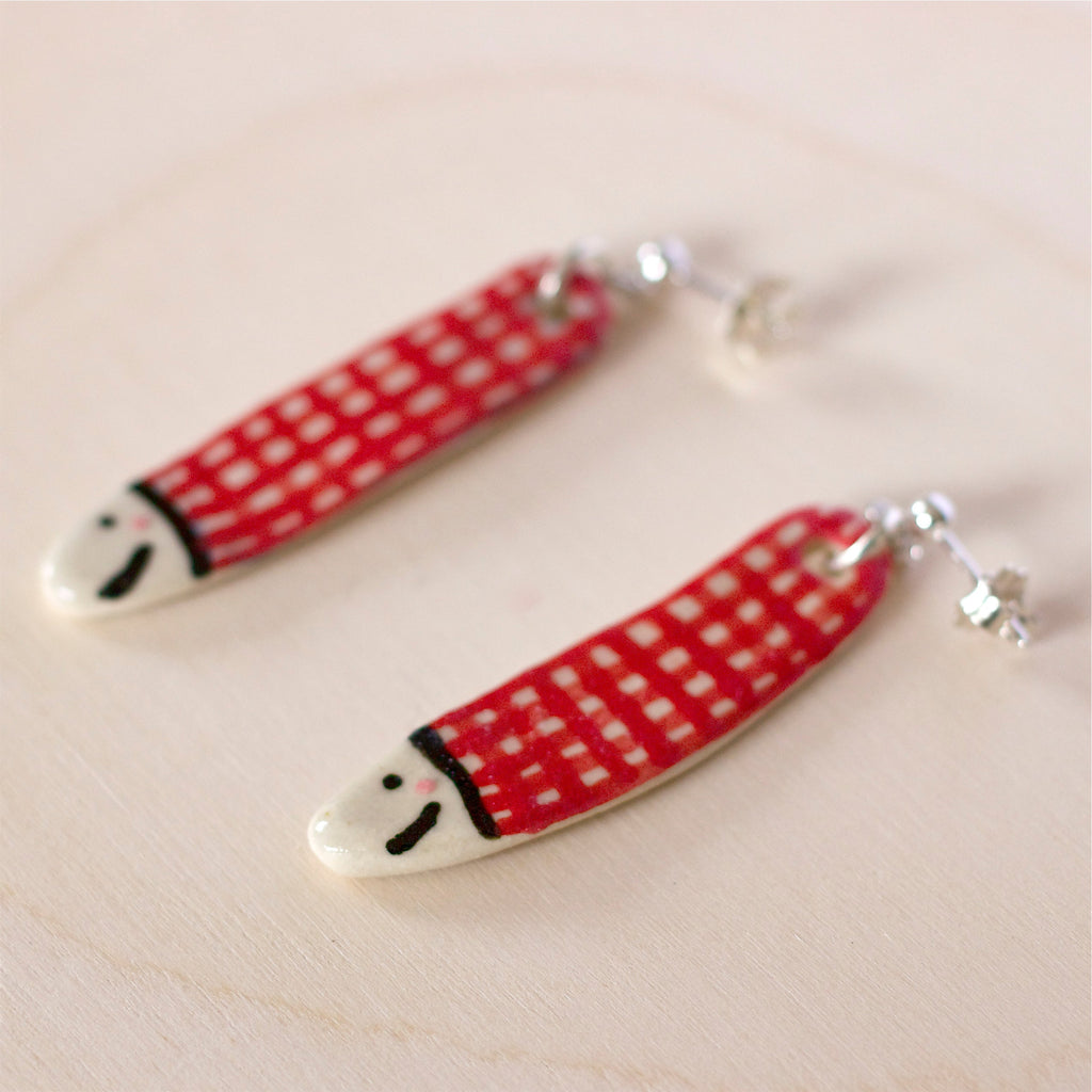 Pair of handmade ceramic earrings shaped like fish with a red check pattern and small white faces, finished with silver stud backings. Designed by shuh.
