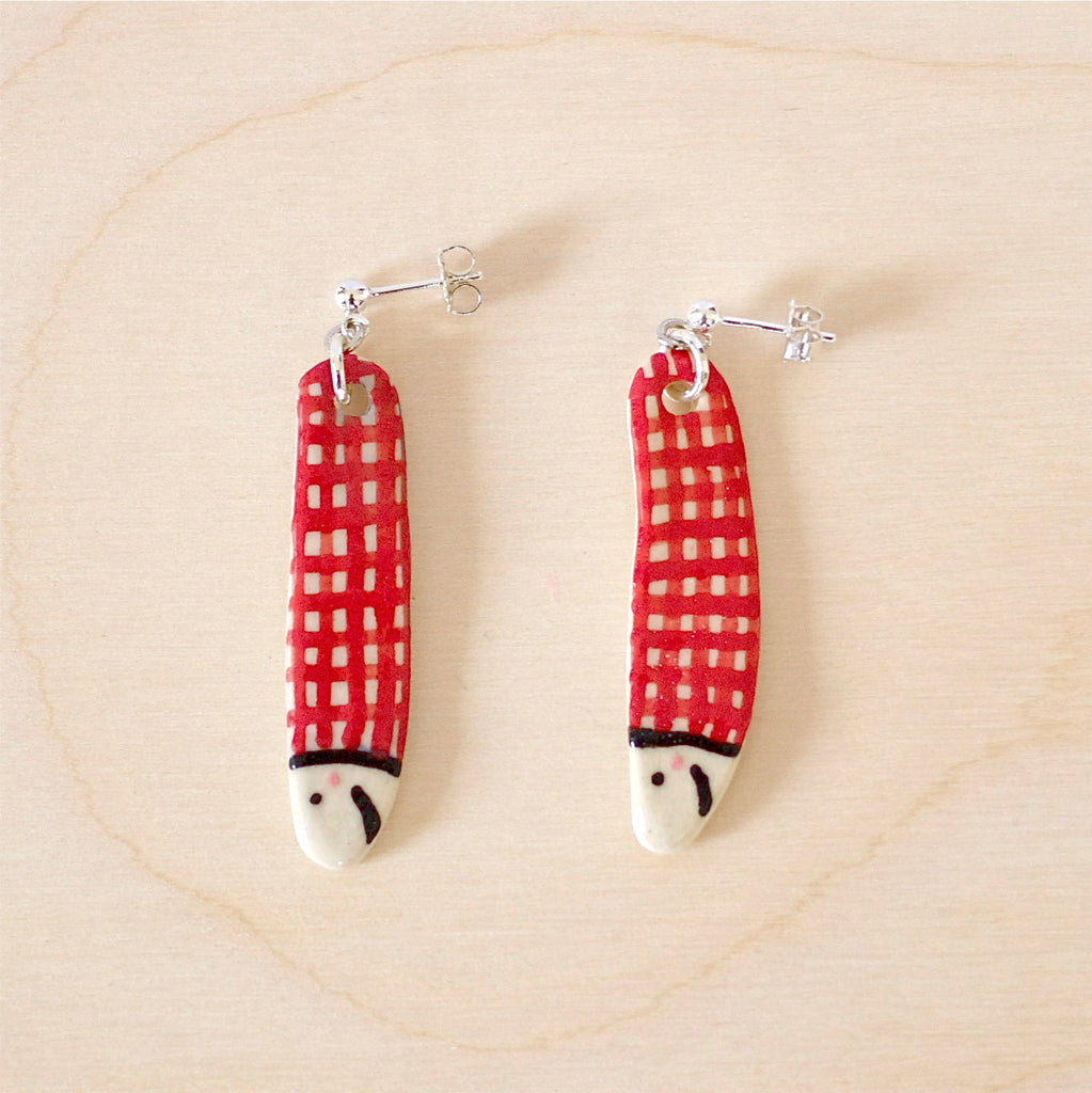 Pair of handmade ceramic earrings shaped like fish with a red check pattern and small white faces, finished with silver stud backings. Designed by shuh.