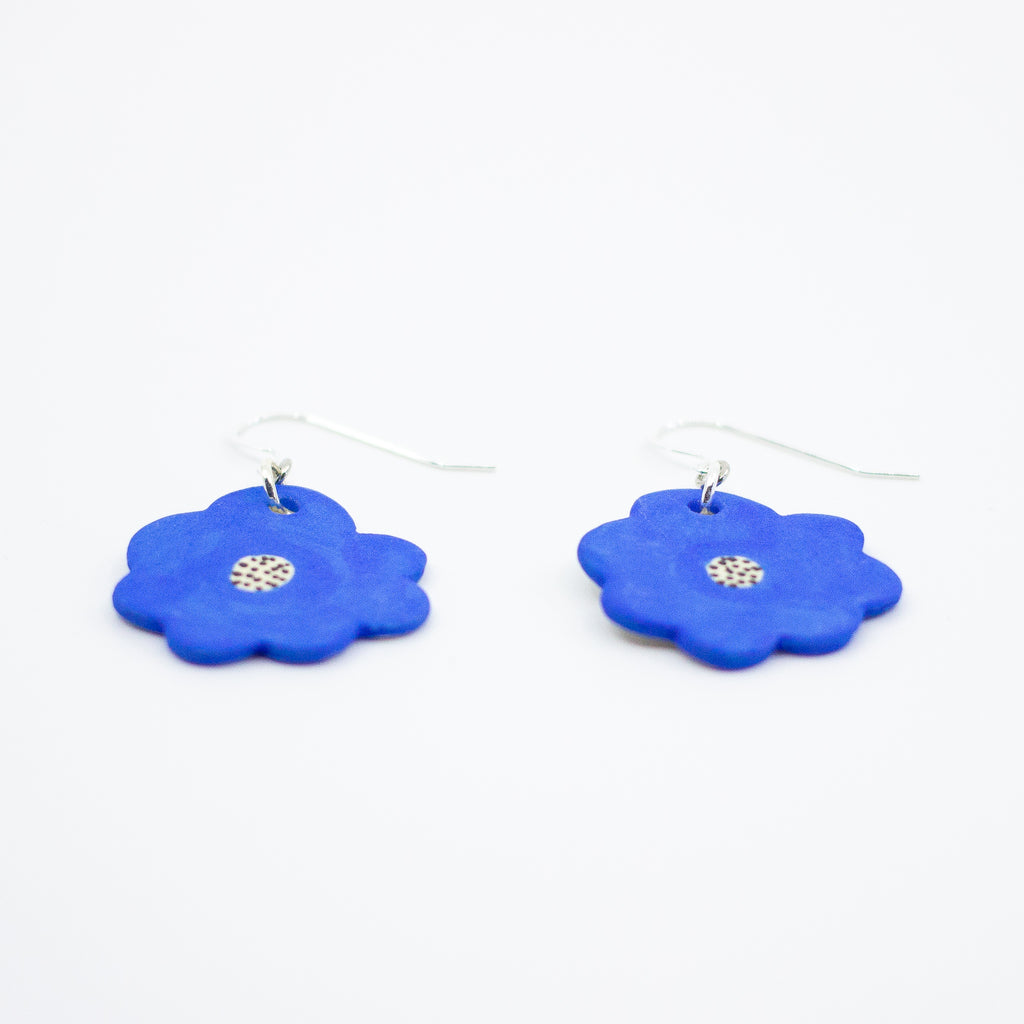 Flat view of a pair of handmade ceramic earrings shaped like bright blue flowers, each with a speckled cream and black centre. Finished with silver hooks.