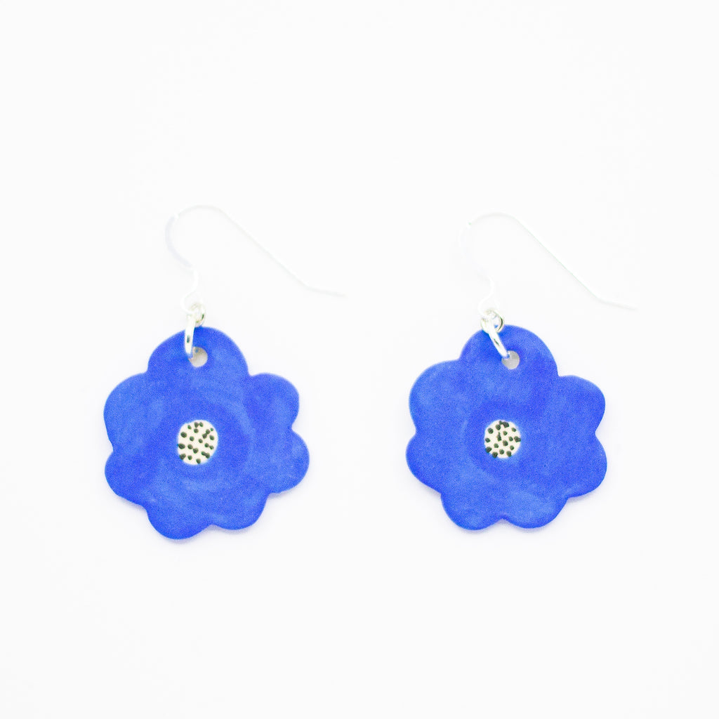A pair of handmade ceramic earrings shaped like bright blue flowers, each with a speckled cream and black centre. Finished with silver hooks.