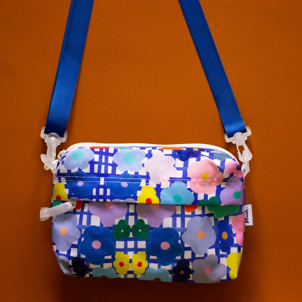 Front view of the flower everyday bag. Alternate blue strap. Bright flower illustrations over a blue and white checkered background.