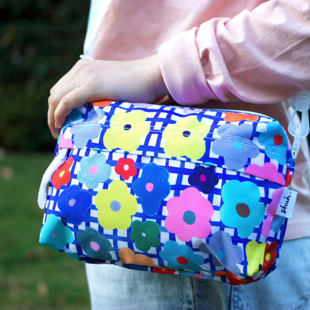 The flower everyday bag being carried over a shoulder. Bright flower illustrations over a blue and white checkered background.
