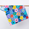 The flower tea towel, hanging from a rack. Bright multicoloured flowers on a blue and white chequered background. 