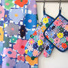 A front view of the Flower tea towel, oven mitt, and pot holder. Multicoloured flowers over a white background with blue stripes. 