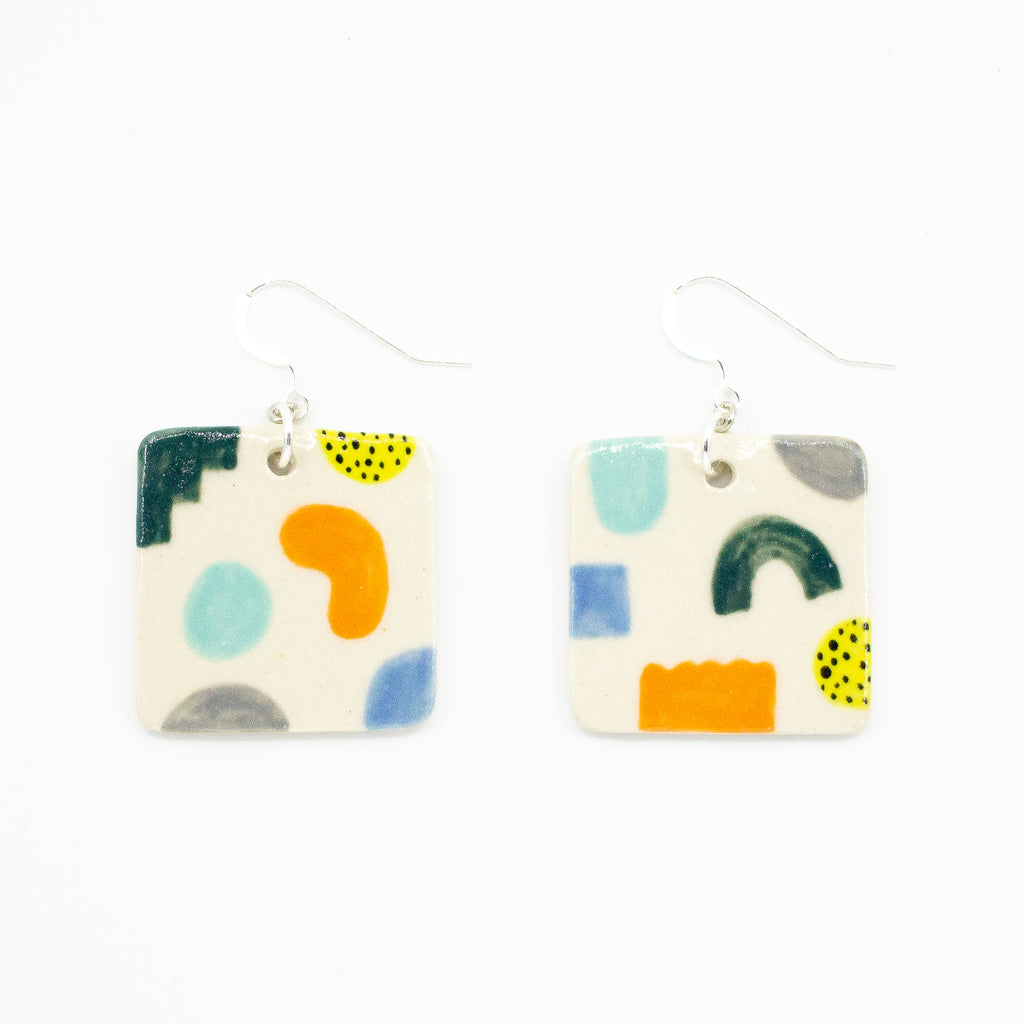 A pair of handmade ceramic earrings with square-shaped tiles featuring abstract colourful shapes in orange, blue, yellow, green, and black. Finished with silver hooks for pierced ears.