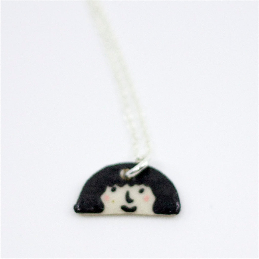Closeup view of a Sterling silver necklace featuring a hand-painted ceramic pendant with a playful face and fuzzy hairstyle, displayed on a white background.