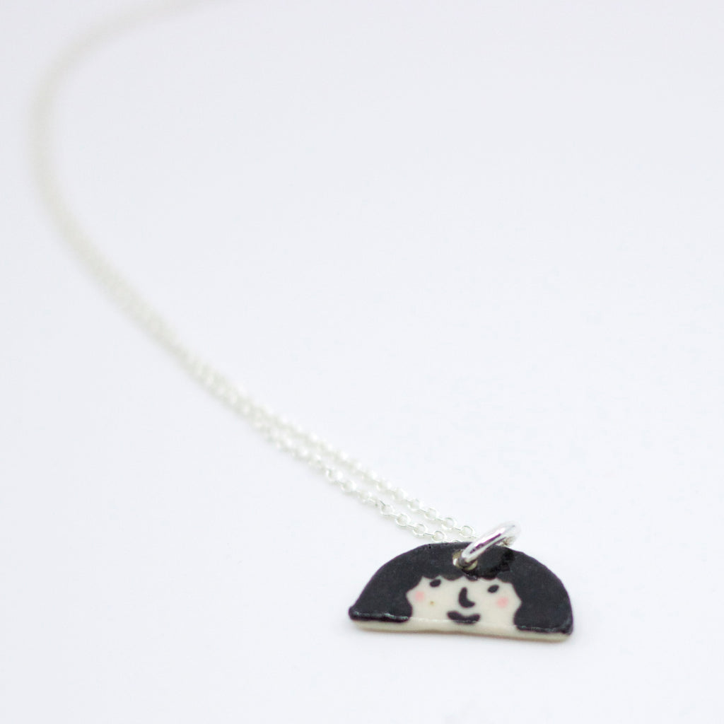 Sterling silver necklace featuring a hand-painted ceramic pendant with a playful face and fuzzy hairstyle, displayed on a white background.