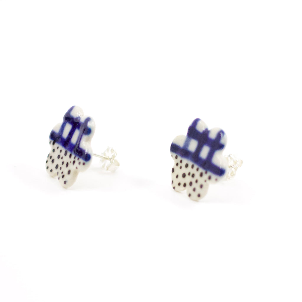 Side view of a pair of handmade ceramic stud earrings in a flower-like shape with indigo check pattern on top and black speckles on the bottom. Sterling silver studs. Designed by shuh.