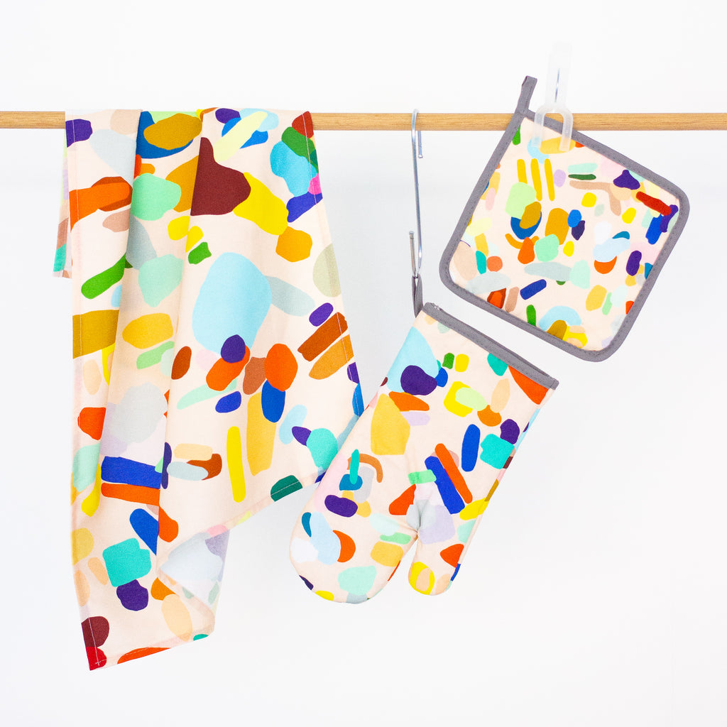 The Hanging Out tea towel, oven mitt, and pot holder front view, hanging from a rack. Multicoloured abstract design on a peach background. 