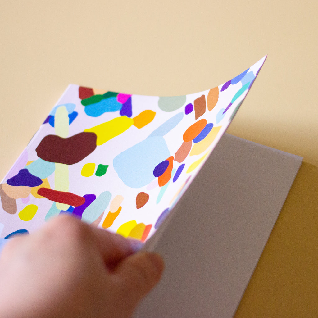Another front view of the Hanging Out notebook. Cover features multicoloured shapes on a light background. Open to show blank pages inside. 