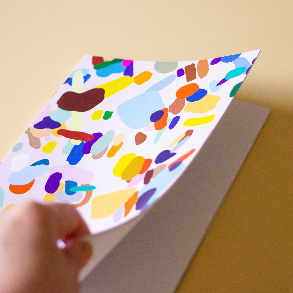 Front view of the Hanging Out notebook. Cover features multicoloured shapes on a light background. Open to show blank pages inside. 