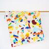 The Hanging Out tea towel, front view, hanging from a rack. Multicoloured abstract design on a peach background. 