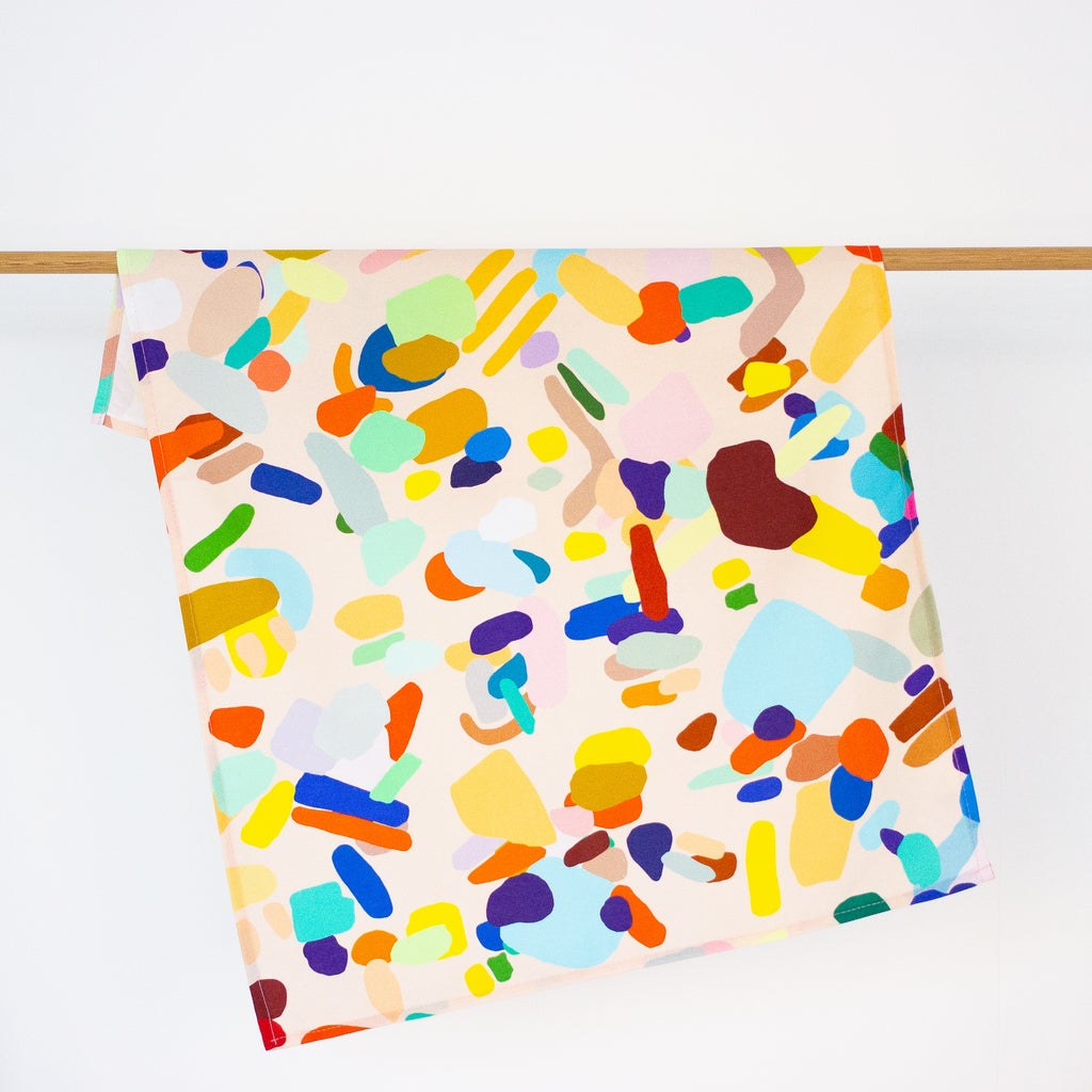 The Hanging Out tea towel, front view, hanging from a rack. Multicoloured abstract design on a peach background. 