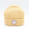 Front view of a tan wide knit beanie with a folded brim, featuring a smiling embroidered flower patch on the front, displayed against a plain white background.