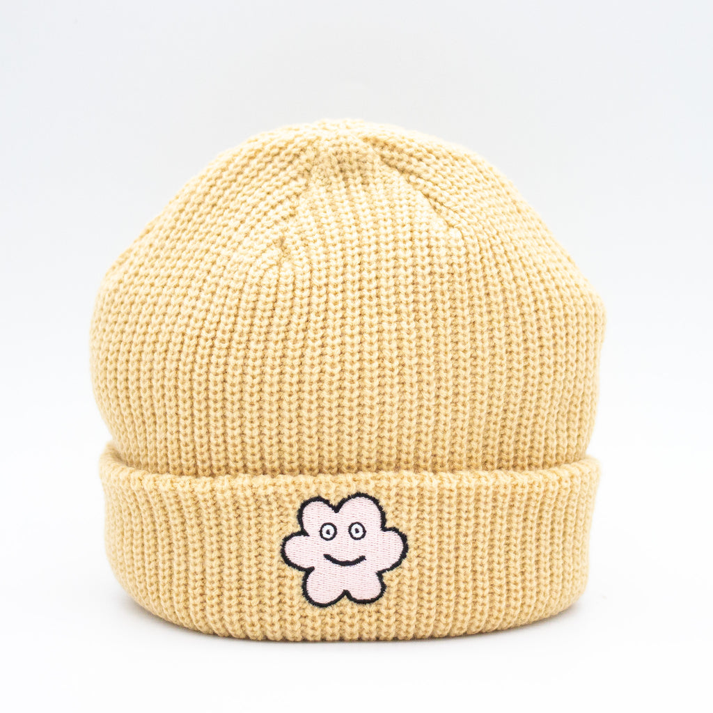 Front view of a tan wide knit beanie with a folded brim, featuring a smiling embroidered flower patch on the front, displayed against a plain white background.