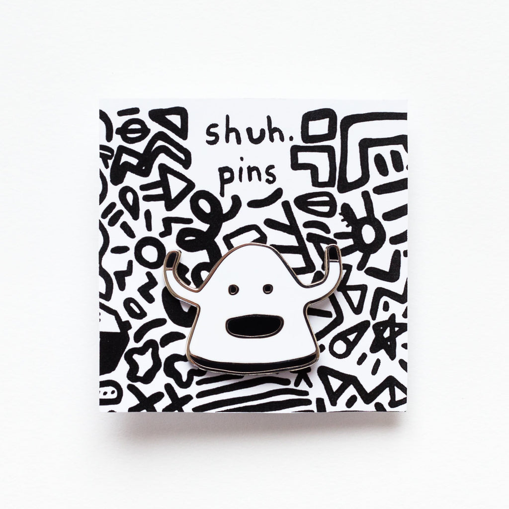 Enamel pin featuring a hand-drawn happy monster character with a big smile and bold black outline, on a card with squiggle designs and 'shuh. pins' labelling. 