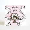 Enamel pin featuring a hand-drawn face wearing a cat hat, displayed on a pink backing card with illustrated cats and “shuh. pins” branding.