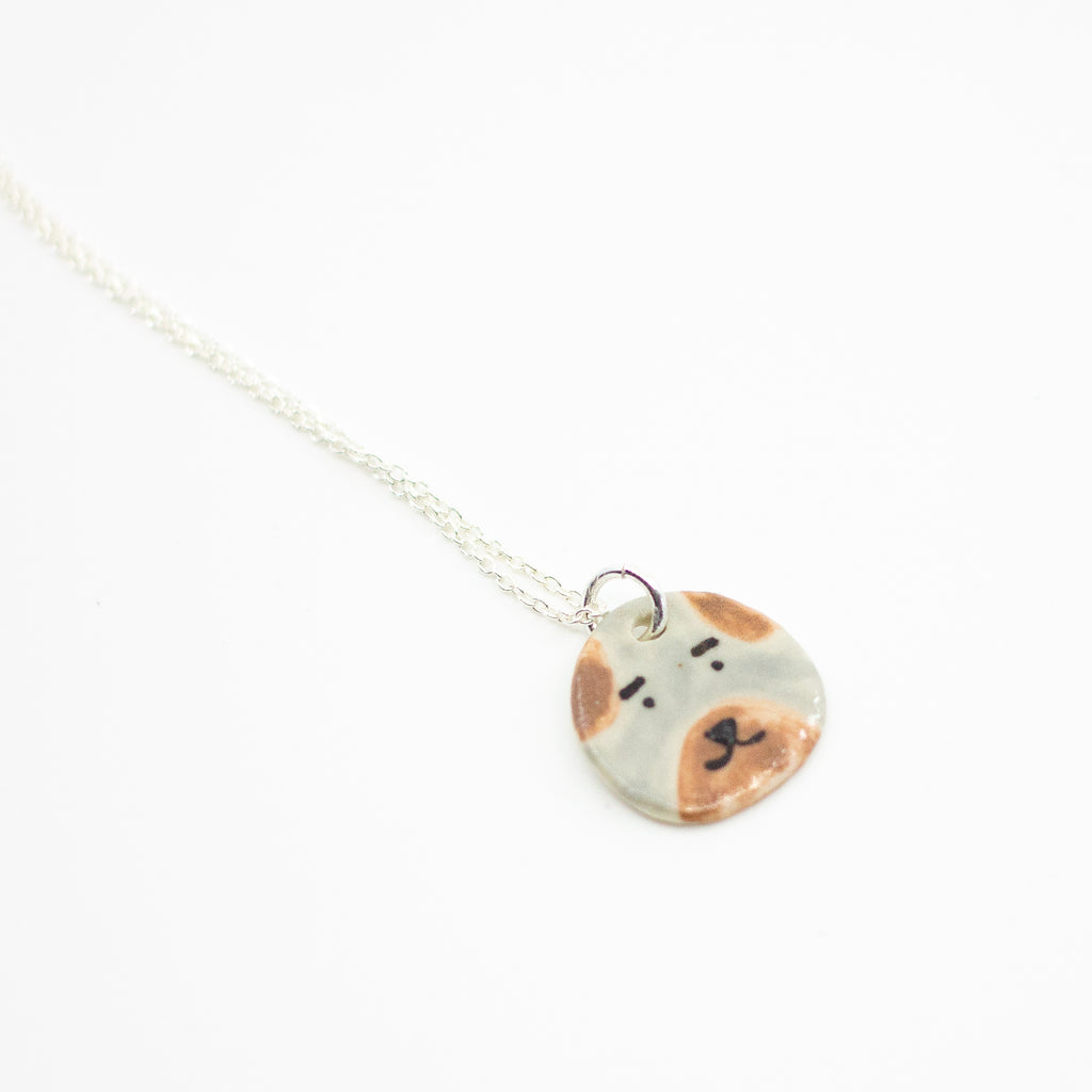 Sterling silver necklace featuring a round ceramic pendant hand-painted with a dog’s face in beige and brown tones, displayed on a white background.