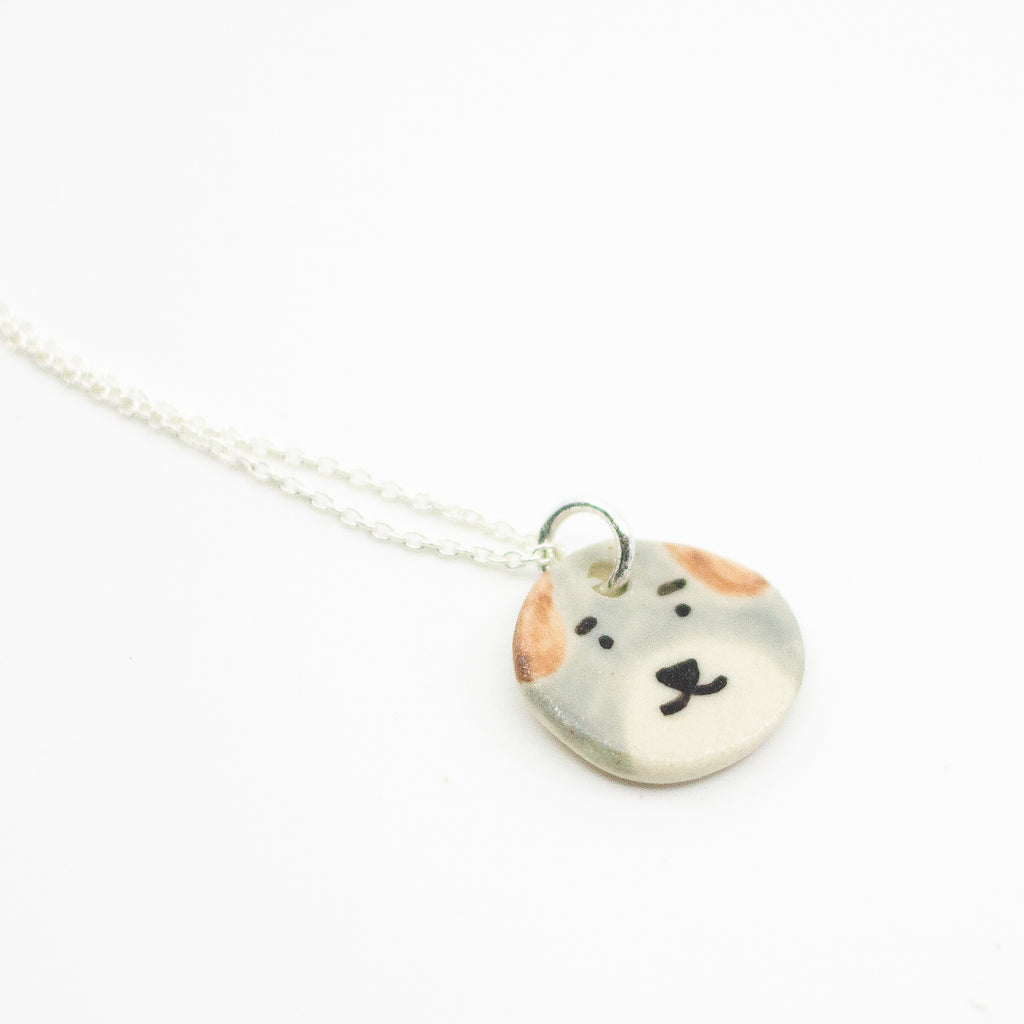 Sterling silver necklace featuring a round ceramic pendant hand-painted with a light grey and beige dog face, including brown ears and a small black nose and mouth, displayed on a white background.