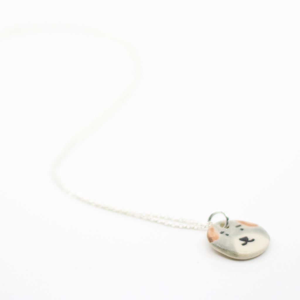 Sterling silver necklace featuring a round ceramic pendant hand-painted with a light grey and beige dog face, including brown ears and a small black nose and mouth, displayed on a white background.