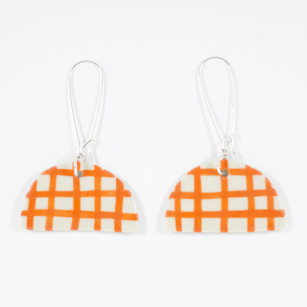 Two ceramic half moon shaped earrings with sterling silver hooks. Ceramic pendants are half circles with orange and white gingham pattern, hand painted. 