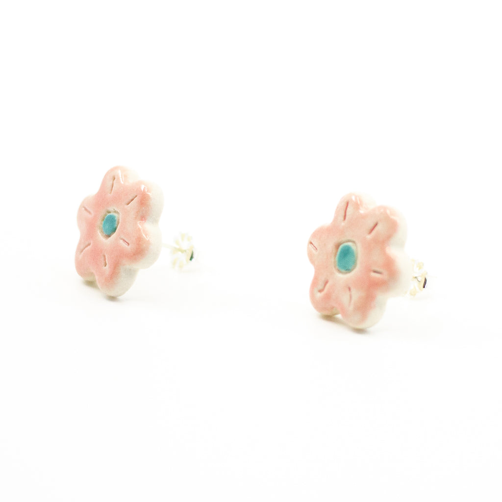 Side view of a pair of handmade ceramic stud earrings shaped like pink flowers, featuring carved petal details and a teal blue centre. Finished with Sterling Silver posts.