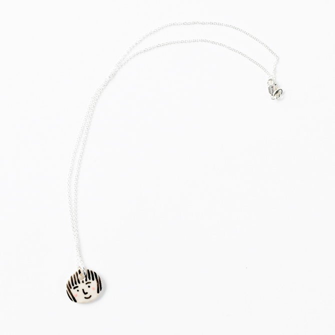 Sterling silver necklace with a small, round hand-painted ceramic pendant featuring a playful girl’s face illustration, displayed on a white background.