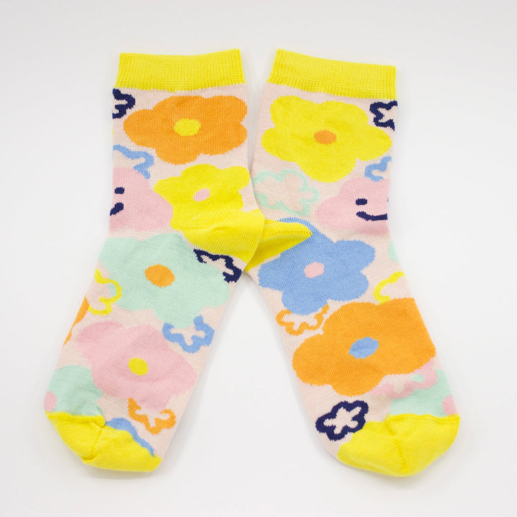 Front view of the sunny bunch socks. This vibrant design features hand-drawn flowers in bright, cheerful hues, blooming in shades of yellow, orange, pink, and blue. Happy and warm smiles peek through the design, reminding you to embrace joy in every season. With a sunny yellow cuff, heel, and toe, these socks bring a warm, uplifting energy to any outfit.