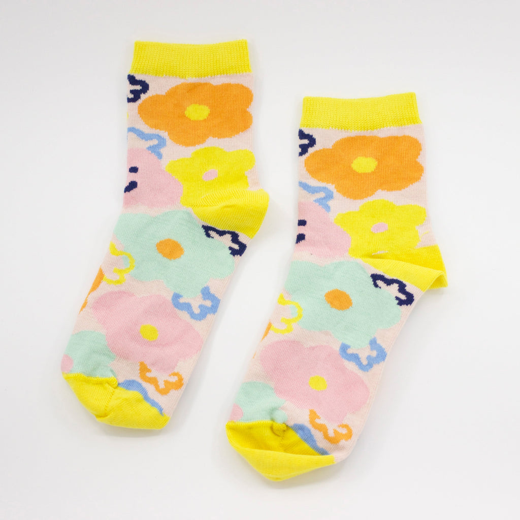 Side by side view of the sunny bunch socks. This vibrant design features hand-drawn flowers in bright, cheerful hues, blooming in shades of yellow, orange, pink, and blue. Happy and warm smiles peek through the design, reminding you to embrace joy in every season. With a sunny yellow cuff, heel, and toe, these socks bring a warm, uplifting energy to any outfit.