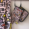 Front view of the sweet oven mitt, tea towel, and pot holder. Street inspired design, pink, black, and yellow designs on a pink background. 