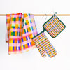 The Tartan tea towel, pot holder, and oven mitt, all hanging from a rack. Pink, green, orange, white, purple, blue layers in a tartan design. 