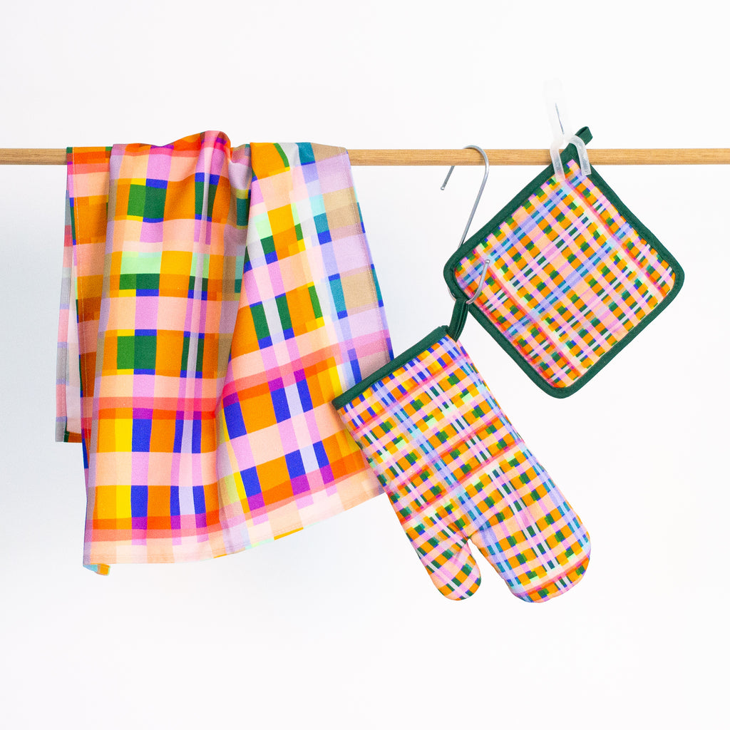 The Tartan tea towel, pot holder, and oven mitt, all hanging from a rack. Pink, green, orange, white, purple, blue layers in a tartan design. 