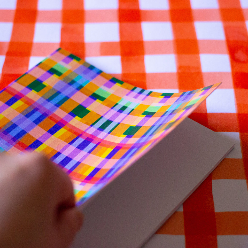 Front view of the Tartan notebook, with multicoloured tartan print. Purple, blue, green, yellow, and orange. Open to show white blank pages inside. 