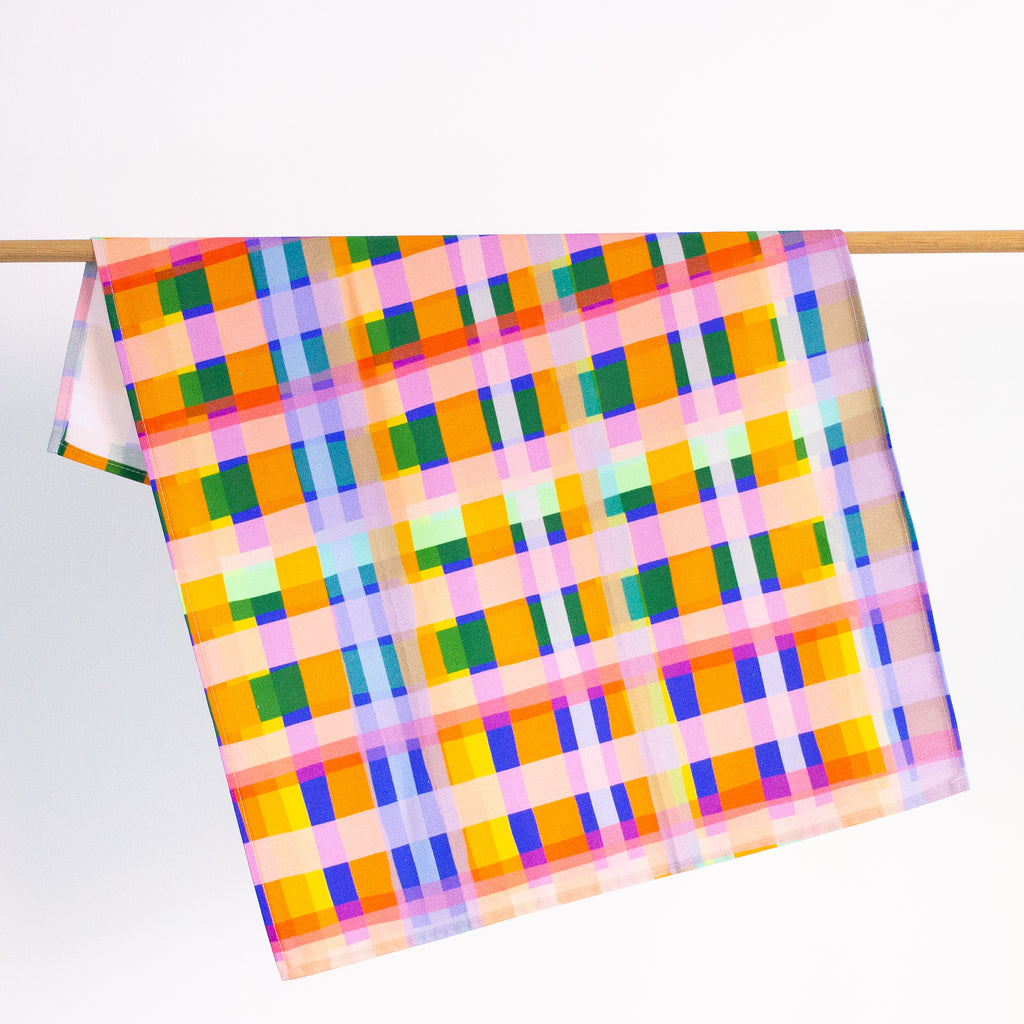 The Tartan tea towel hanging from a rack. Pink, green, orange, white, purple, blue layers in a tartan design. 