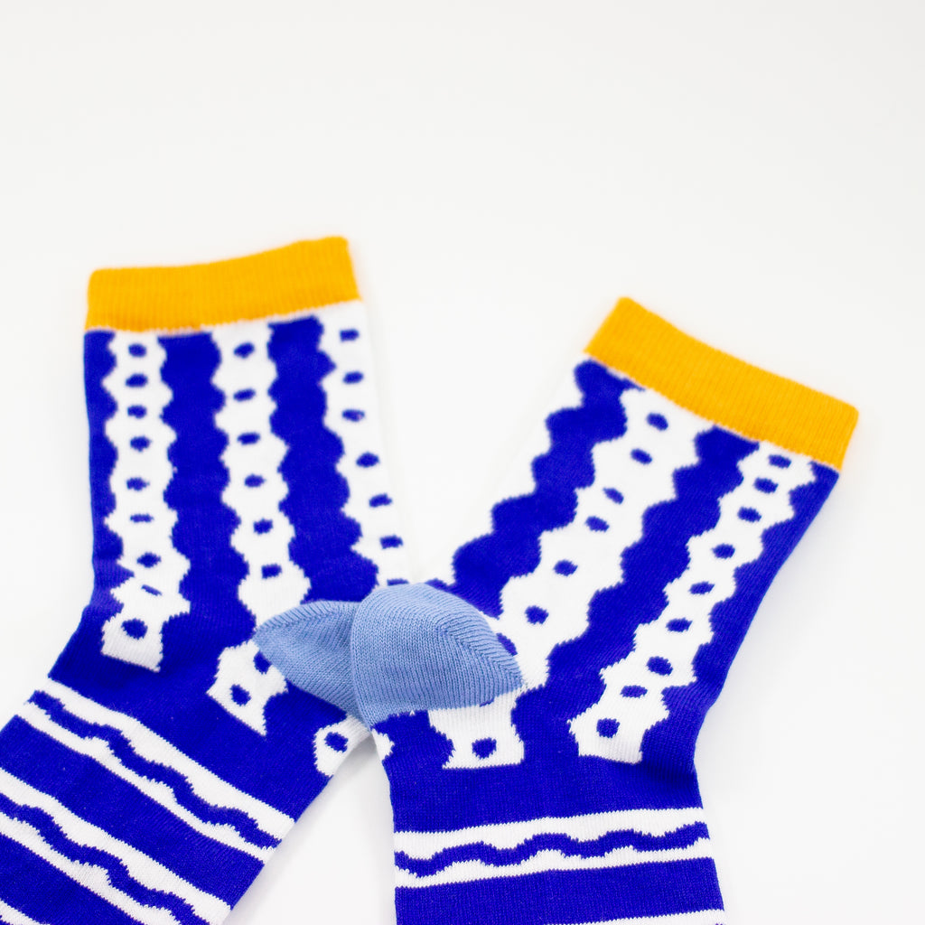 Closeup of the cuff of the TENGU socks. Featuring a bold blue and white pattern of playful waves, dots, and stripes, these socks bring energy and excitement to every step. The bright yellow cuff and toe add a pop of colour, while the soft blue heel keeps things stylish and unique.